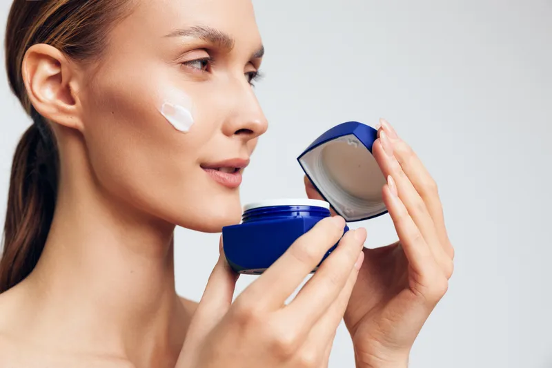 Close-up of a woman applying white cream on her cheek, open blue jar beside on light background, showcasing skincare routine and facial care with soft studio lighting
