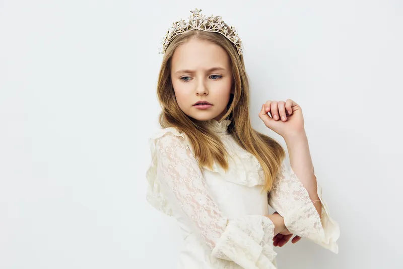 A pensive young girl wearing a lace dress and a crown, showcasing elegance and innocence against a soft, minimalistic background, perfect for portraits or fashion imagery
