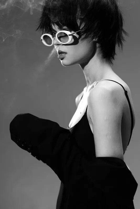 Black and white portrait of a stylish woman wearing glasses and smoke near her lips with a moody atmosphere.