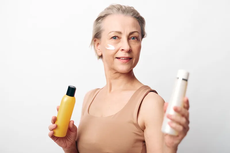 Mature woman with short hair applies facial cream holding skincare bottles on white background. Concept of beauty routine, self-care, and healthy skin for older adults.