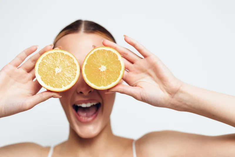 Woman smiling holding citrus orange and lemon halves like glasses, hands framing her face, fresh vitamin rich fruit, clean studio background, playful healthy lifestyle