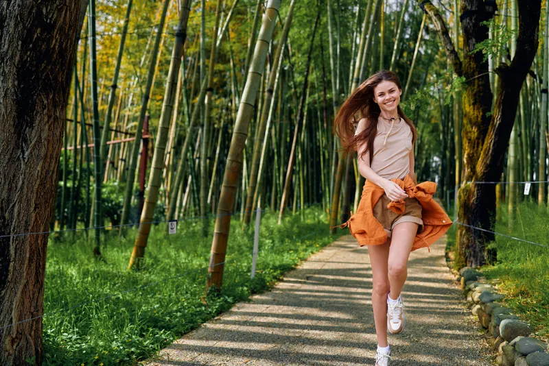 Woman running along a bamboo path in a sunlit forest, smiling and carefree. Girl in casual skirt and jacket captured in motion on a leafy trail, summer outdoor lifestyle portrait.