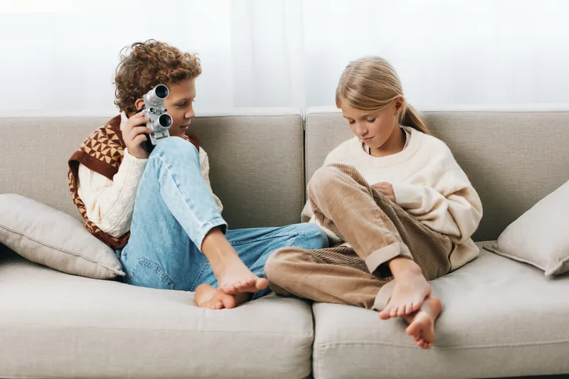 Two children relax on a light sofa; one boy aims a vintage camera while the other girl sits nearby, wrapped in a cozy blanket, sharing a warm, playful home moment.