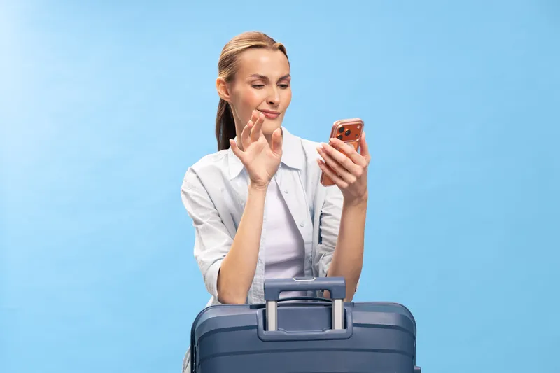 Young woman with ponytail waves hand while video calling on smartphone. She stands behind blue suitcase on light blue background. Concept of travel, communication, and remote connection.