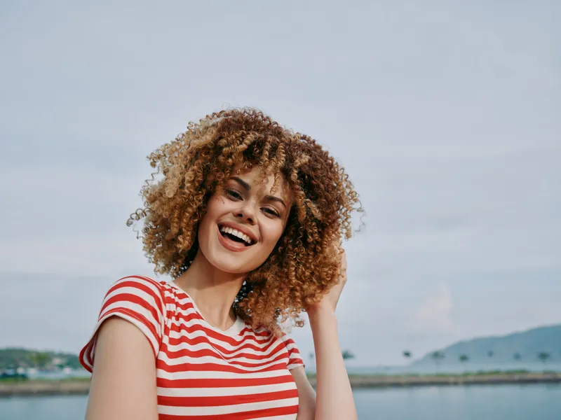 Portrait of a joyful woman with curly hair and a red striped tee, standing outdoors by a calm shoreline with a bright sky and gentle breeze, vibrant colors, natural light, and a candid smile.