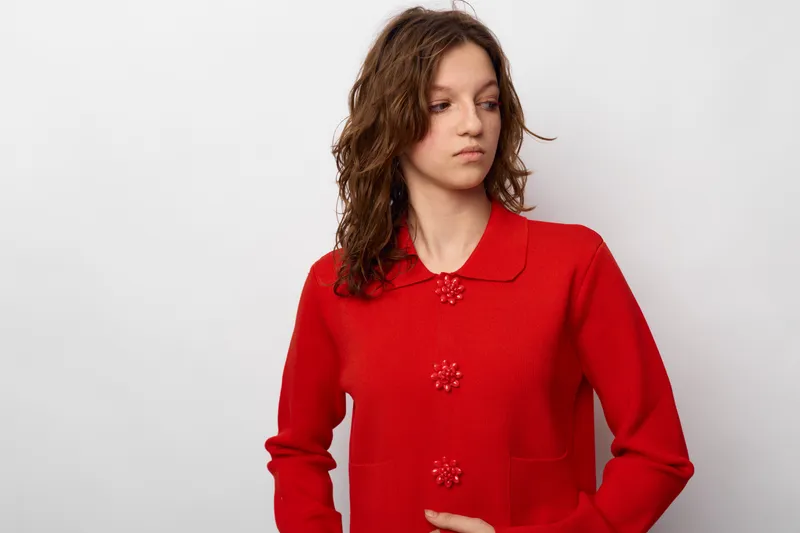 Young woman in red sweater looks away with serious expression on white background. Casual fashion portrait with natural light and minimalistic style. Concept of contemplation and mood.