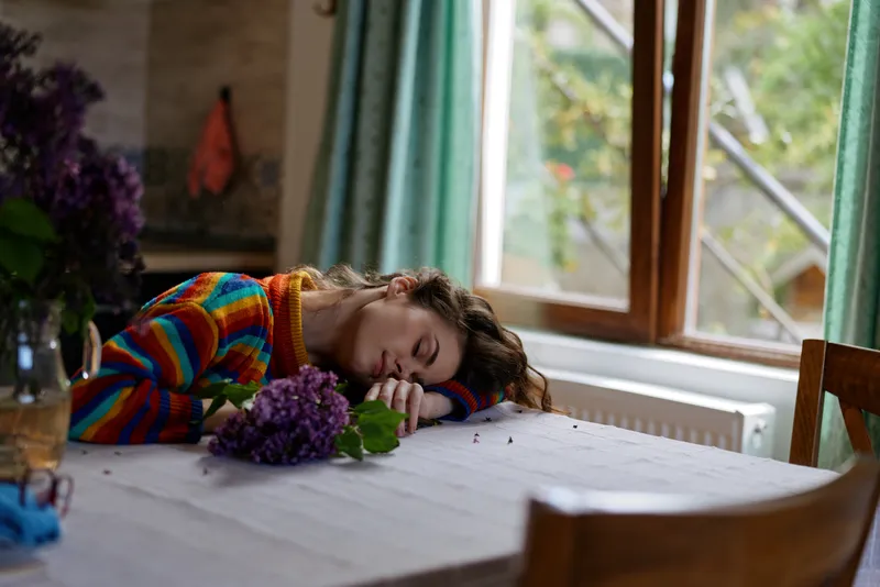 Young woman in a colorful sweater rests her head on a dining table by a sunlit window, surrounded by purple flowers, creating a peaceful intimate home moment.
