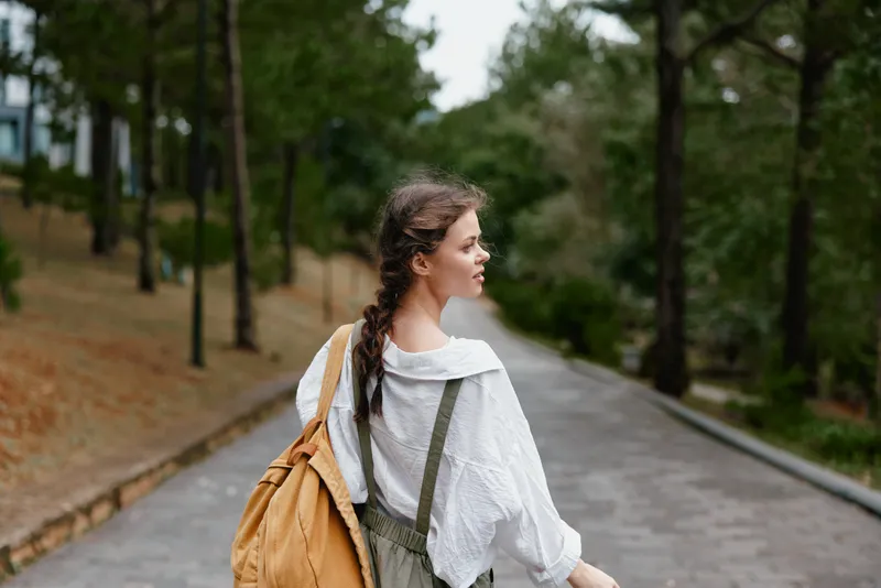 Young woman walking in a serene park, wearing a relaxed outfit with a backpack, showcasing a sense of freedom, nature, and adventure