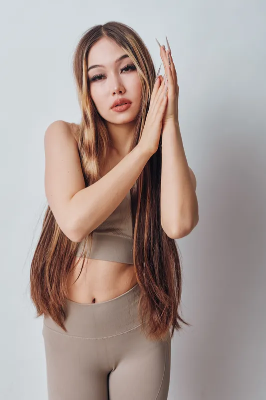 Young woman with long straight hair wearing beige crop top and pants posing with clasped hands in front of light background in studio. Beauty, fashion, casual outfit, portrait.
