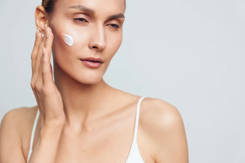 Young woman applying moisturizer on face with gentle hand movement, smooth skin, natural makeup, white background, skincare routine, beauty concept closeup