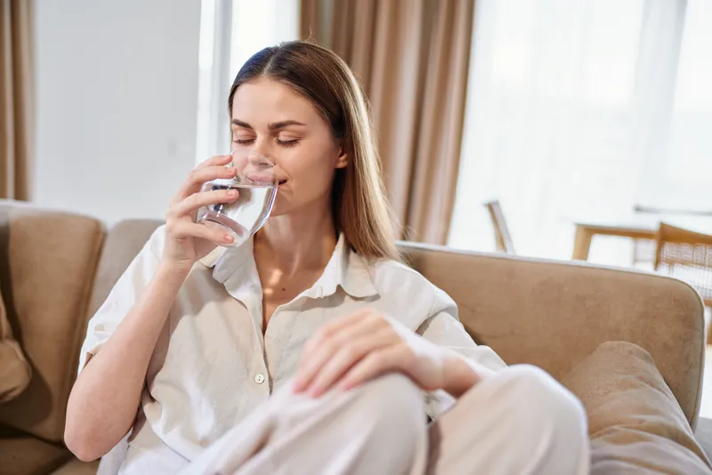 Woman drinking water from a glass on a couch in a bright home living room, relaxation and wellness moment, healthy hydration, casual clothing and peaceful indoor lifestyle.