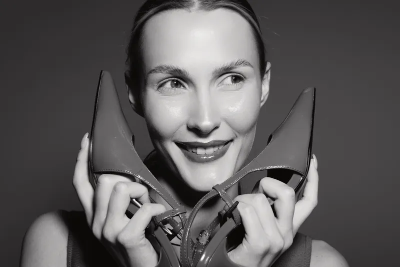 Smiling white woman with light skin holding red high heels near face, looking aside with happy expression in studio. Concept of fashion, style, and footwear.