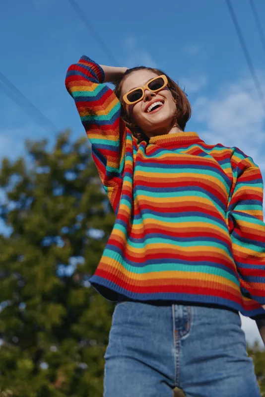 Woman smiling in a colorful striped sweater and yellow sunglasses, young adult in her 20s wearing a casual outfit, leaning back and laughing outdoors under blue sky for lifestyle fashion.