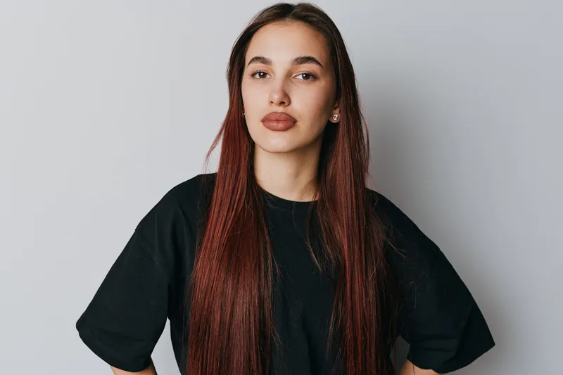 Woman with long straight hair and casual black t shirt standing with hands on hips looking confident against plain background in studio portrait.