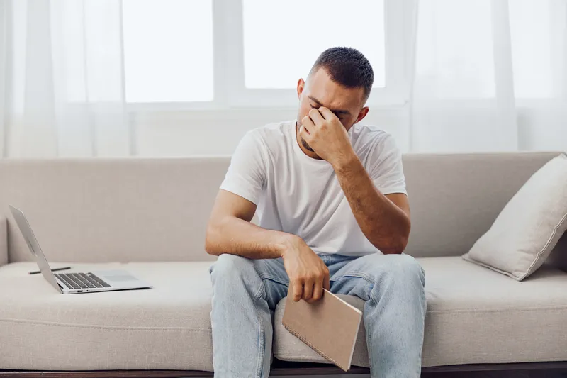 Stressed man sitting on a couch, holding his forehead with frustration. A laptop is nearby, showcasing a modern and minimalist home office setting.