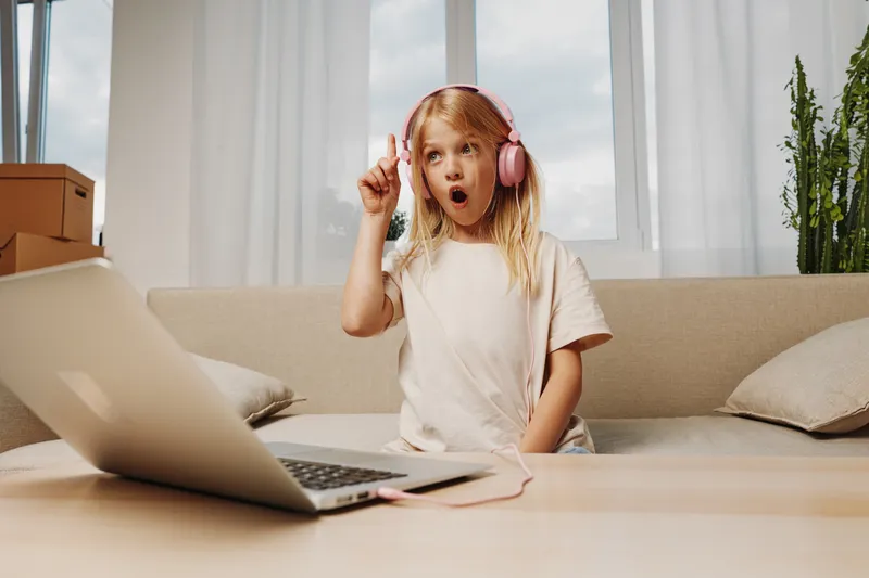 Excited young girl with blonde hair wearing pink headphones watches laptop on sofa in bright living room, expressing surprise and joy. Concept of remote learning, entertainment, and childhood