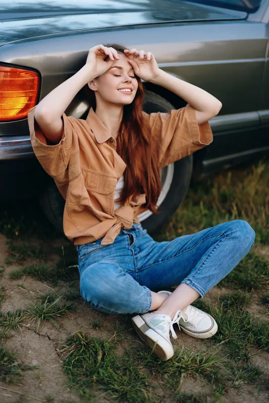 Young woman enjoying the outdoors by a vintage car, wearing a casual brown shirt, relaxed with a joyful expression, beautiful natural setting.