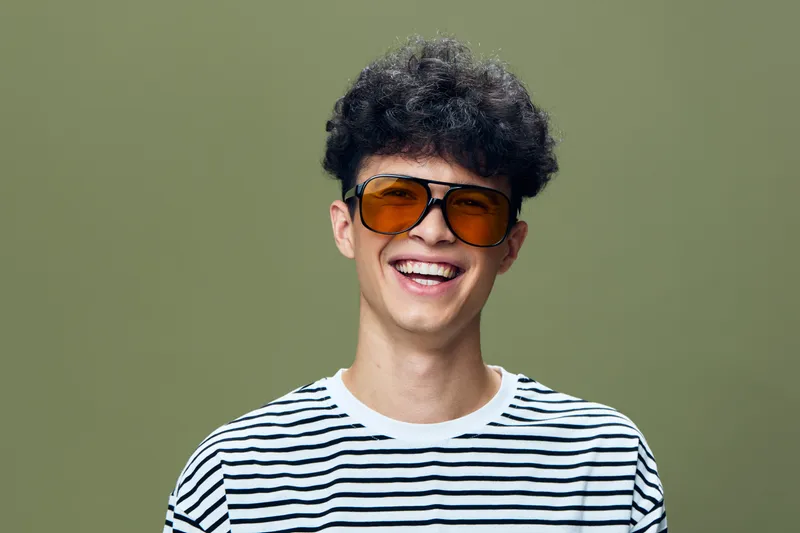 young man smiling wearing sunglasses and striped t-shirt with curly hair against green background, casual and cheerful portrait of a happy male