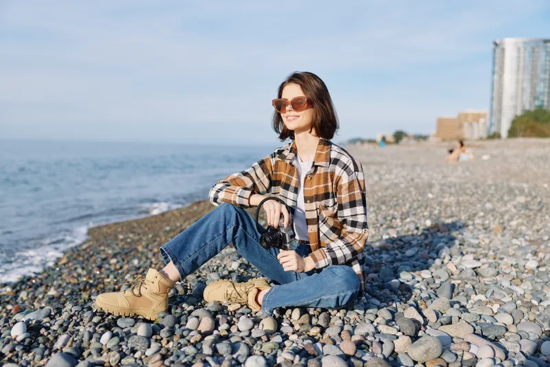 Casual photographer sitting on a pebble beach with a camera, wearing a plaid shirt, jeans and sunglasses, enjoying coastal light and a relaxed mood as waves roll in, distant buildings on the horizon,