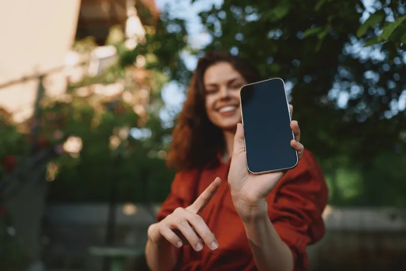 A cheerful woman in casual wear holds a smartphone outdoors, smiling at the camera in a lifestyle moment