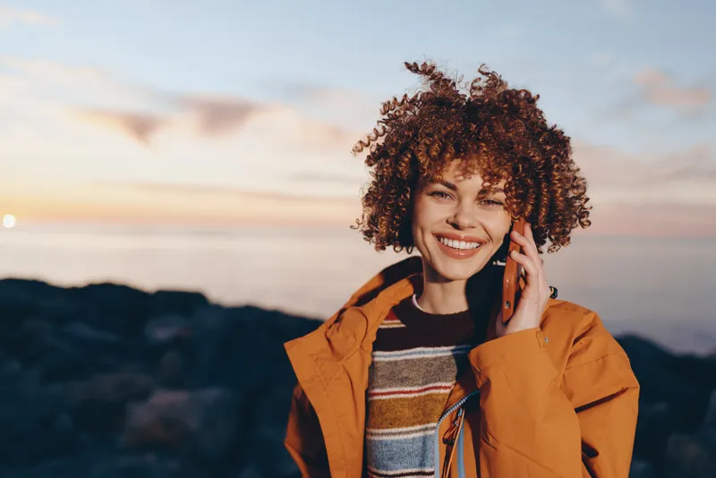 Smiling woman with curly hair outdoors, speaking on a phone at sunset. Warm jacket, relaxed lifestyle, mountain landscape background. Natural light for travel happy mood today.