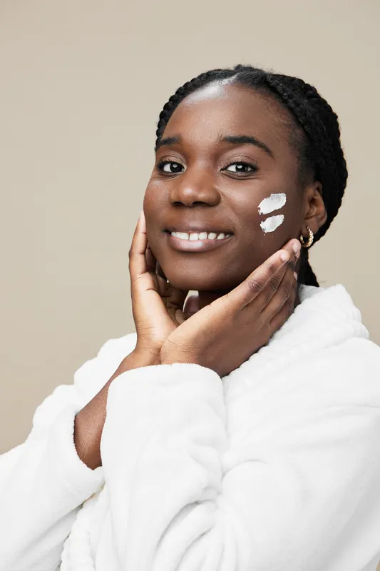 Beautiful young Black woman applying skincare cream on her face, smiling gently, wearing a white robe against a light beige background, promoting self care and skincare routines