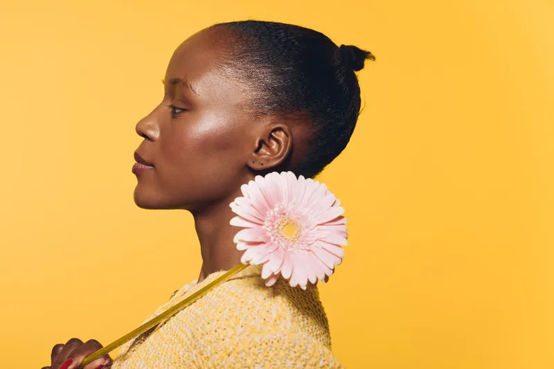 Profile of a young black woman with dark skin holding a pink flower on yellow background. Concept of natural beauty, elegance, and floral aesthetic with soft lighting and calm expression.