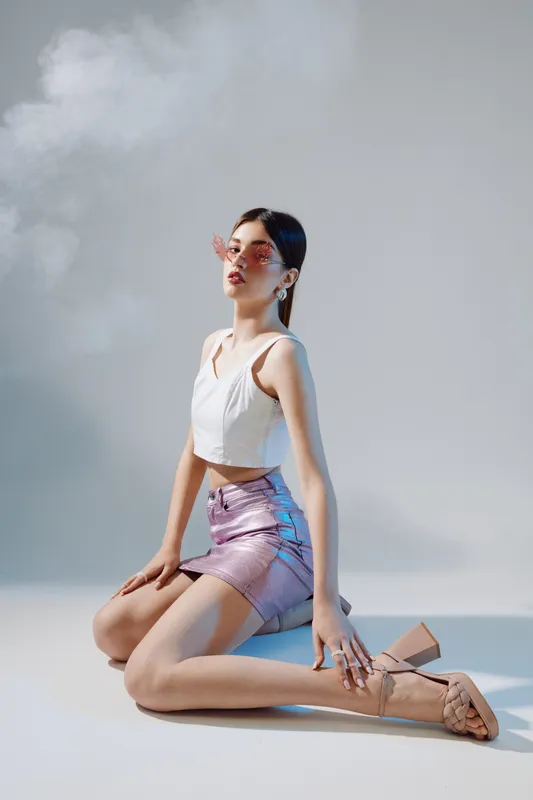 Young woman in stylish fashion outfit kneels on the floor wearing a white top and purple skirt. Soft lighting highlights her beauty and confident pose in a minimal studio setting.