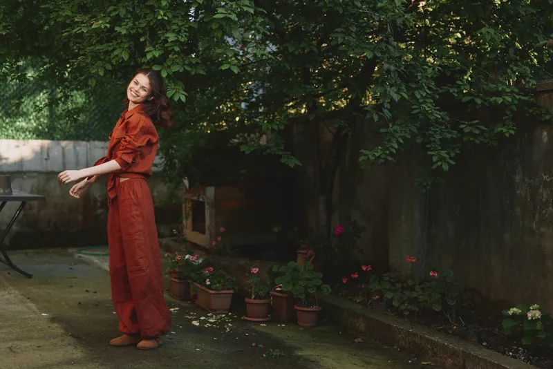 Young woman in a rust red jumpsuit circles playfully in a garden courtyard, smiling softly as potted plants line the path and dappled light filters through trees