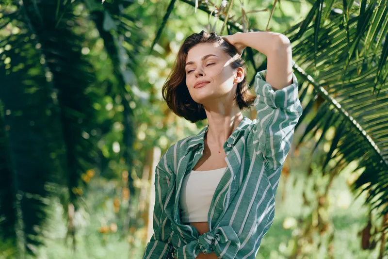 Portrait of a relaxed woman enjoying a sunny day outdoors, wearing a striped shirt and white top amid lush greenery.