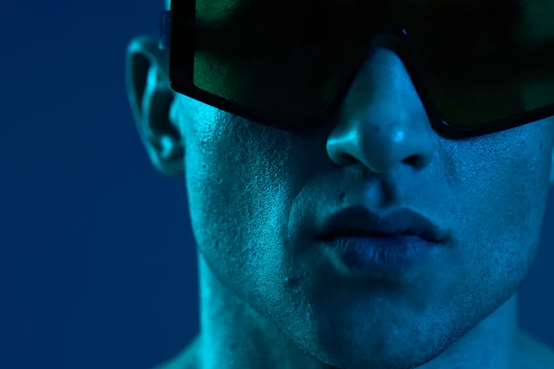 Close-up of a man wearing stylish sunglasses, captured in blue light, highlighting his facial features and creating a modern atmosphere in a minimalistic setting.