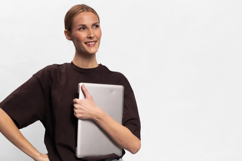 Smiling young woman holding a laptop close to her chest stands against a plain white background. Casual outfit and confident pose create a professional yet relaxed mood.