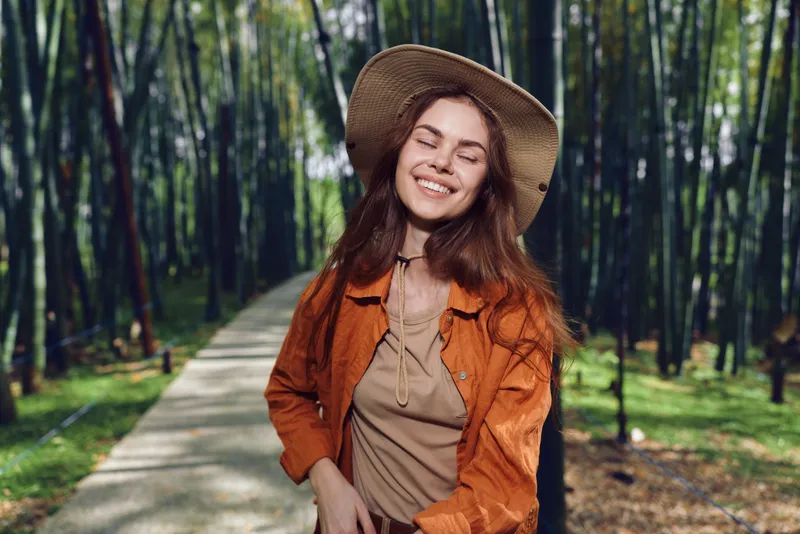 Woman smiling in a straw hat on a sunlit park pathway through bamboo forest, portrait of a happy nature lover in casual orange jacket, outdoors relaxation, travel and joyful expression.