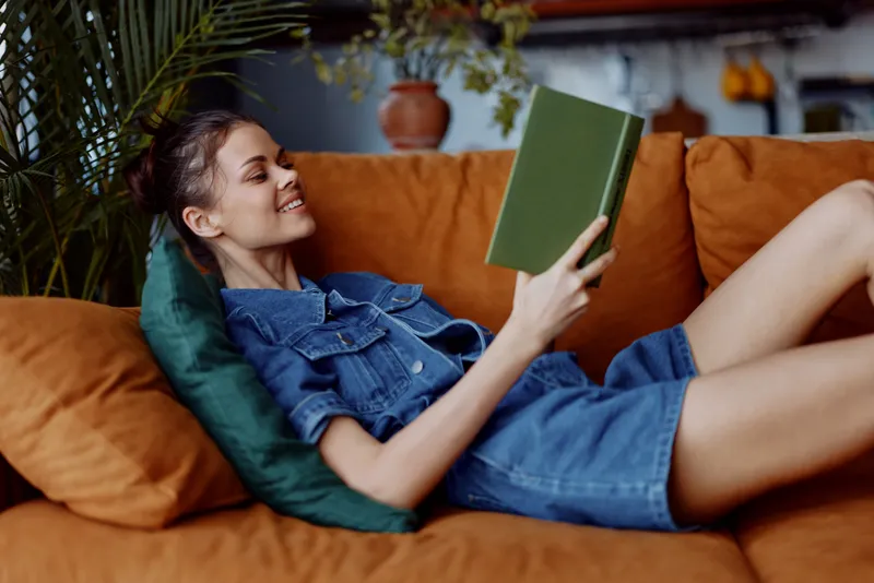 Женщина отдыхает на диване в уютной комнате, читая книгу; на заднем плане видно растение