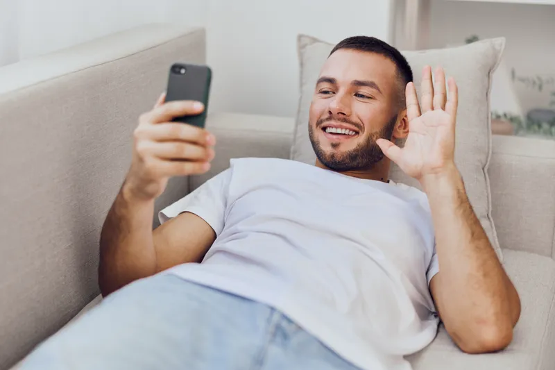 Smiling man enjoying a video call, representing the blend of personal and work life. His casual attire and relaxed position reflect modern communication trends.