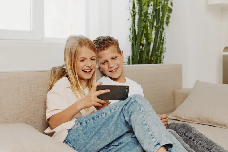 Happy smiling children sitting on couch watching smartphone together in bright living room. Concept for family, technology, leisure, childhood, and digital entertainment.