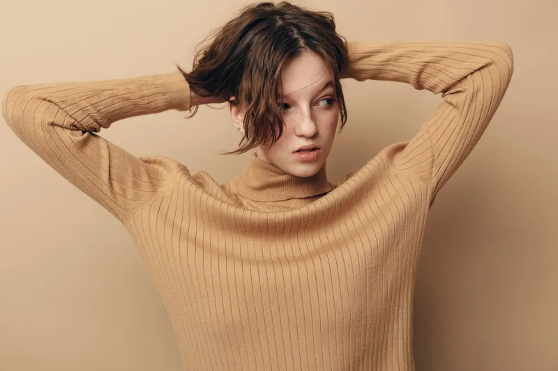Young woman with short curly hair wearing beige sweater looks away with hands behind head. Neutral background creates calm and introspective mood in studio portrait.