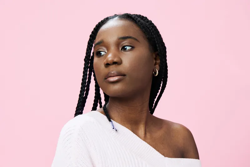 Young Black woman with braids in soft pink background, showcasing thoughtful expression, dressed in pastel sweater, ideal for beauty and lifestyle themes