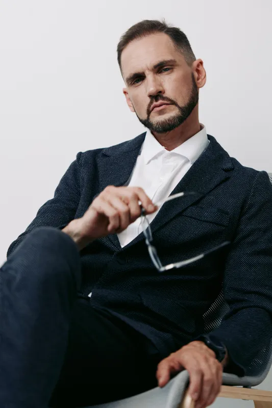 A composed business man in a textured blazer sits against a plain light background, projecting confidence and focus while adjusting his glasses.