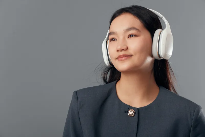 Young woman wearing wireless headphones and looking away with slight smile against plain gray background. Portrait of confident female in dark clothing with natural makeup and long hair for lifestyle