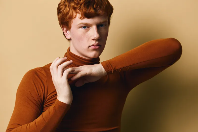 Young man with red hair wearing brown turtleneck sweater posing with arms raised and neutral expression on beige background in studio portrait fashion style.