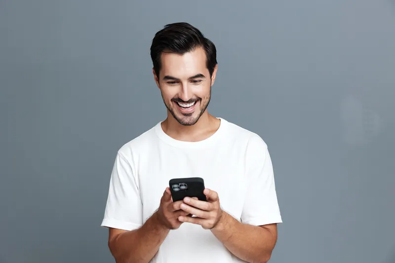 Smiling young man with dark hair wearing a white t-shirt, looking at his mobile phone, isolated on a solid grey background. Modern lifestyle concept.