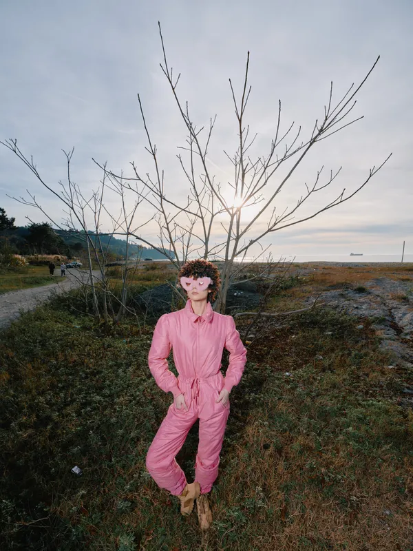 Pink jumpsuit worn by a confident woman in an outdoor portrait beside a bare tree, coastal landscape in soft daylight, cinematic mood, fashion-forward scene for nature and travel themes