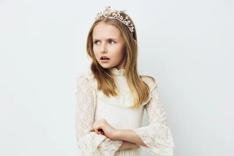 Young girl wearing a crown and a vintage dress, looking surprised against a white background, capturing a whimsical and fairy tale like essence
