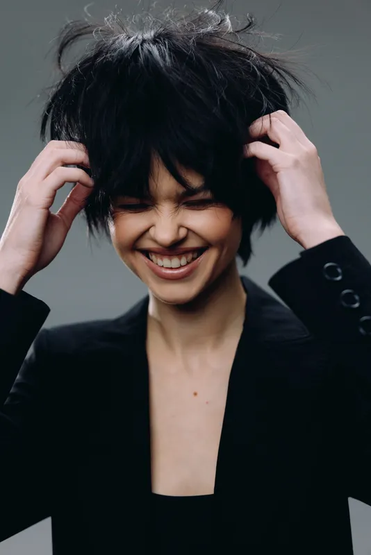 Smiling woman with tousled hair wearing a black blazer, expressing joy and confidence in a simple studio setting.