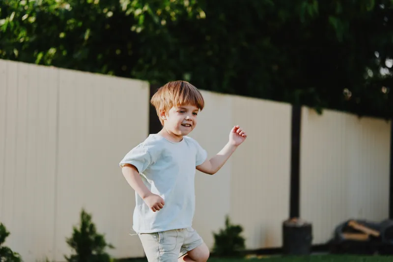 Happy boy running outdoors in a green garden with playful energy, enjoying a sunny day surrounded by nature and a cozy wooden fence.