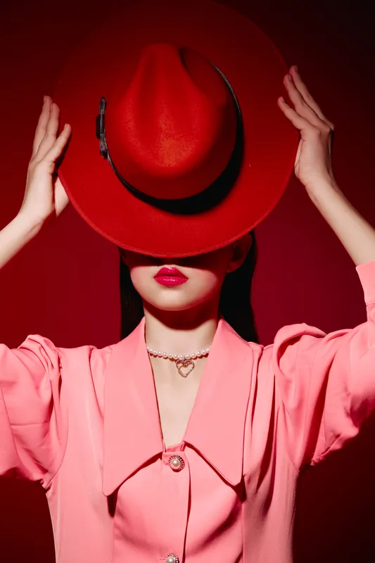 Stylish woman in a pink blouse and red hat holds hat to cover face with bright lipstick under dramatic lighting. Concept of fashion, beauty, and creative portrait.