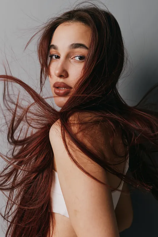 Portrait of young woman with long hair blowing in wind, looking over shoulder with natural makeup and neutral expression against plain background in studio lighting.