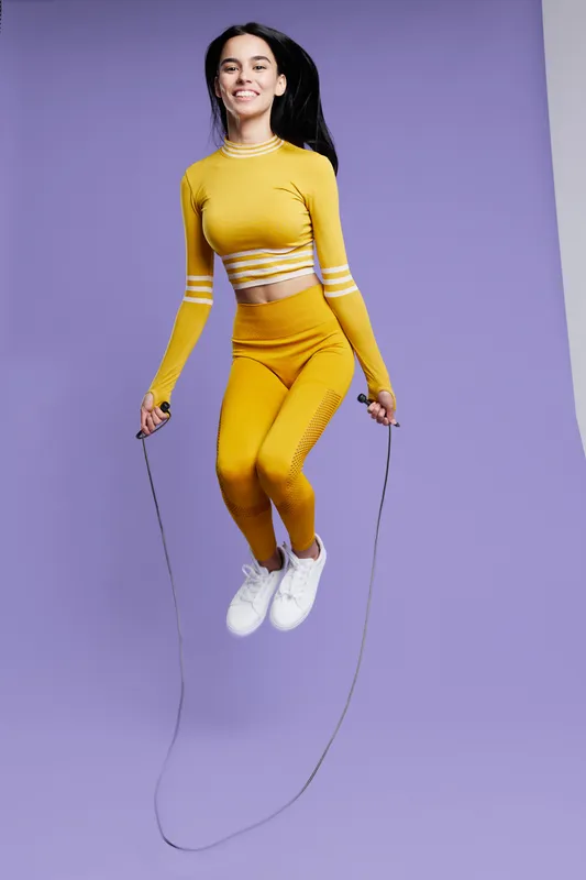 Athletic woman sport fitness jumping rope in yellow activewear, smiling and energetic, isolated on a purple background in dynamic motion.