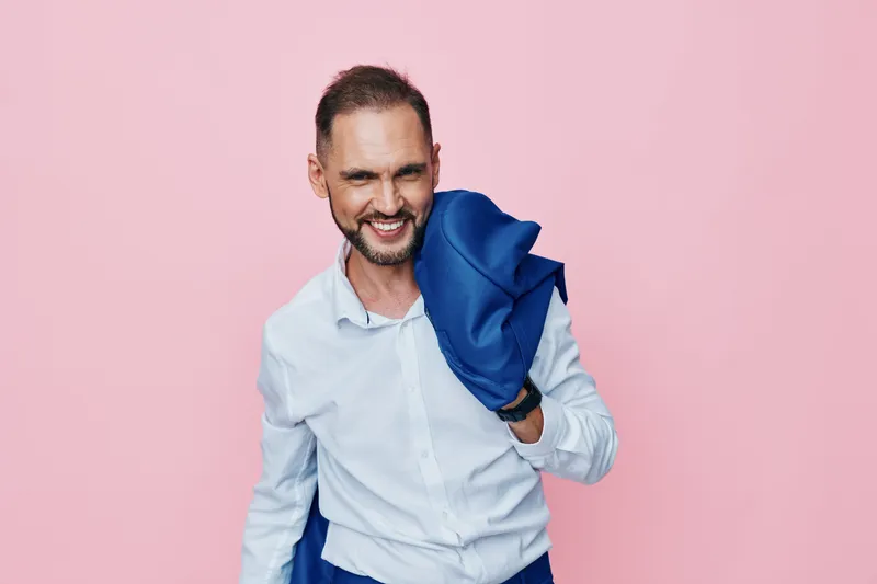 A professional man stands against a vibrant pink background, casually carrying a blue jacket over his shoulder, smiling with confidence and approachable presence.